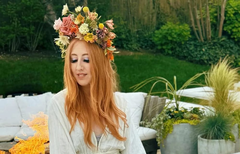 A woman with long rose pink hair in a white dress and massive flower crown looks down and inward. 