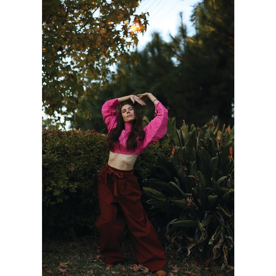 a white woman with long brown hair stretches her arms over her head in a bright pink long sleeved crop top, eyes closed again
